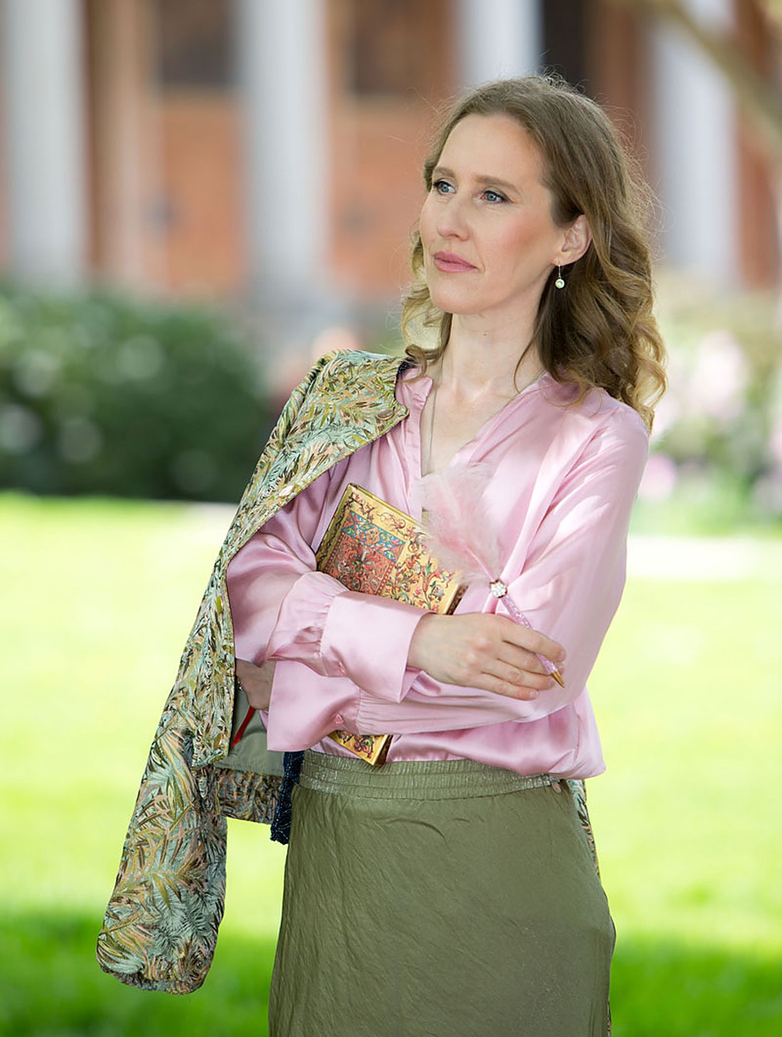 Sylvia Harke mit einem kunstvollen Buch und einer Schreibfeder im Arm, in einer rosa Seidenbluse – Autorin und Muse.